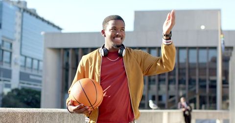African american student holding basketball and waving on urban college campus
