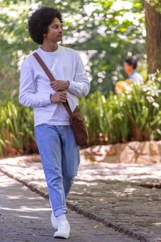 African american man walking through sunlit park carrying crossbody bag, casual style