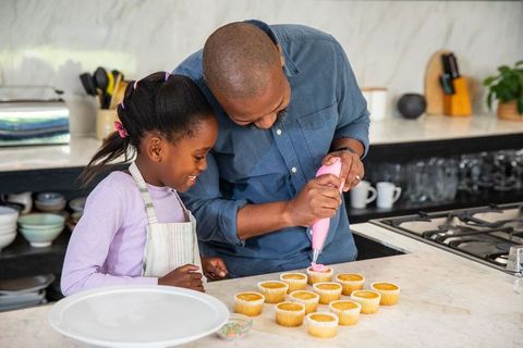 African american father decorating cupcakes with daughter using pink piping bag on counter