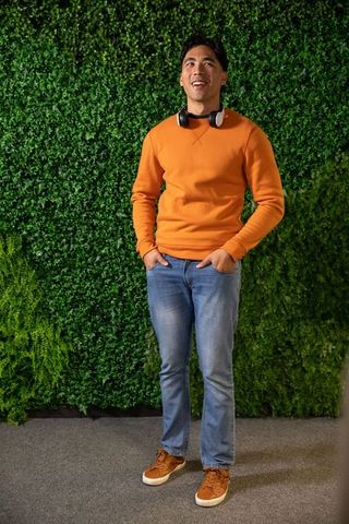 Asian young man standing before green foliage wall wearing orange sweatshirt and headphones