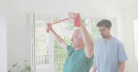 Senior man raising red resistance band while physical therapist assisting at home rehab