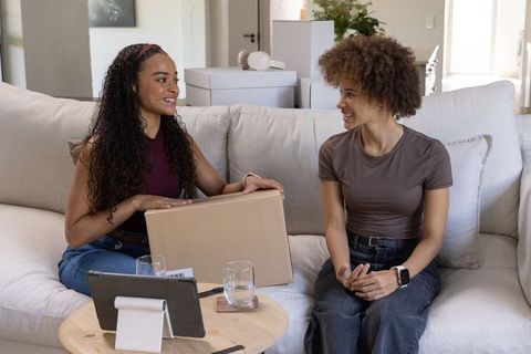 Two african american women unpacking box on sofa, smiling and talking, home interior lifestyle