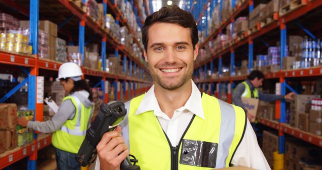 The image shows a smiling warehouse worker holding a scanner inside a large storage facility. The setting features tall shelves filled with boxes and other items. The worker is wearing a high-visibility vest, emphasizing safety and professionalism. Ideal for use in content related to warehouse operations, logistics management, inventory control, and occupational safety. Also suited for promoting teamwork and workplace efficiency in storage and distribution environments.