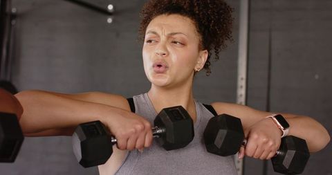 Woman lifting hex dumbbells in gym showing strength and focused intensity, wearing smartwatch