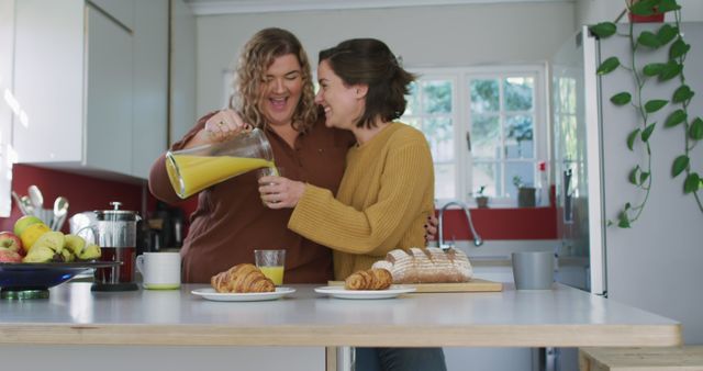 Two friends enjoying breakfast together in a bright, modern kitchen. One is pouring orange juice, while they both smile and laugh. Various fruits, coffee, and pastries are arranged on the counter. Perfect for illustrating friendship, morning routines, and happy moments at home.