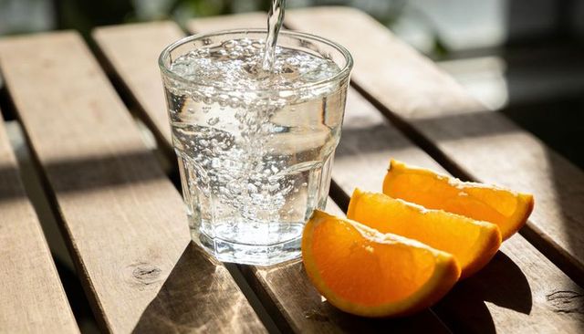 Pouring sparkling water into glass with orange wedges and bubbles on sunlit wooden table