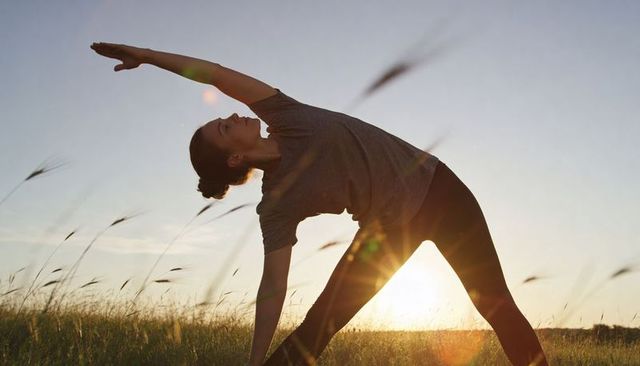 Woman practicing triangle yoga stretch at sunset in meadow with backlight and sun flare