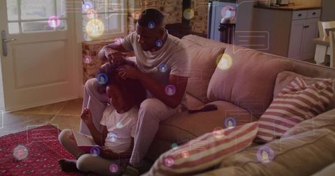 Father braiding daughter's hair while child using tablet in cozy room with digital social bubbles