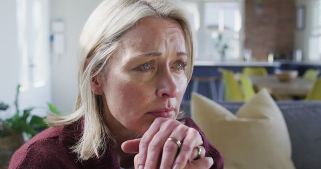 Middle-aged woman with blonde hair looking pensive and worried while sitting at home. The woman appears to be in deep thought or going through an emotional struggle. Useful for articles, blogs, or advertisements focused on mental health, aging, personal struggles, or home living.