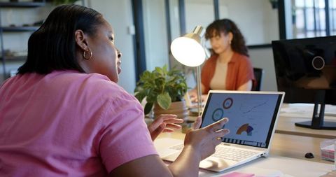 African american woman presenting laptop analytics to colleague in collaborative office