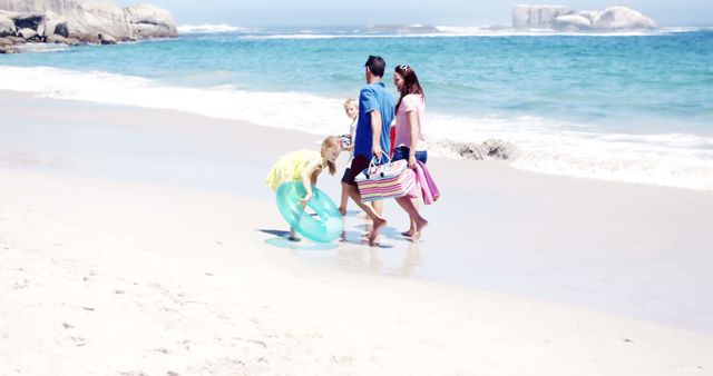 Family with children walking along sandy beach carrying picnic items. Blue ocean waves gently hitting the shore and rocks visible in the distance. Perfect for depicting family vacations, summer activities, or an enjoyable day by the sea.