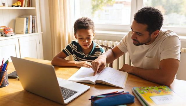 Father helping son with homework using workbook and laptop, focused home learning
