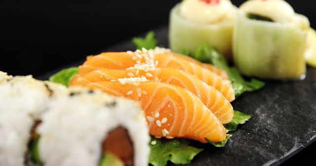 Slices of fresh salmon sashimi are garnished with sesame seeds, accompanied by sushi rolls and cucumber-wrapped delicacies on a slate serving board. Sushi is a traditional Japanese dish celebrated for its delicate flavors and artistic presentation.