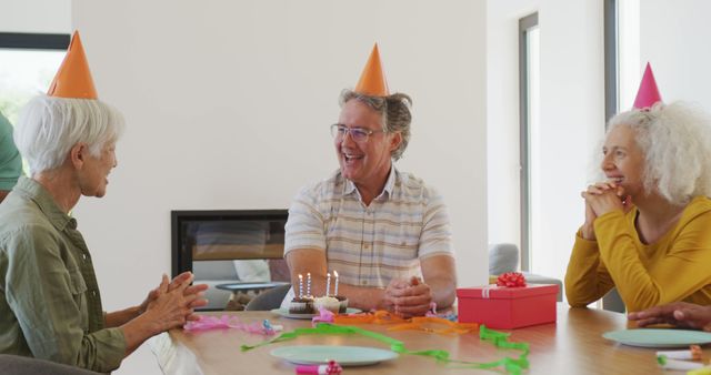 Three senior friends celebrating a birthday at home. They are seated around a table with a cake, candles, gift and colorful decorations. Ideal for concepts related to friendship, celebration, aging, and positivity.