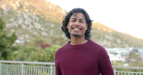 Smiling asian man standing on terrace wearing maroon knit sweater during golden hour