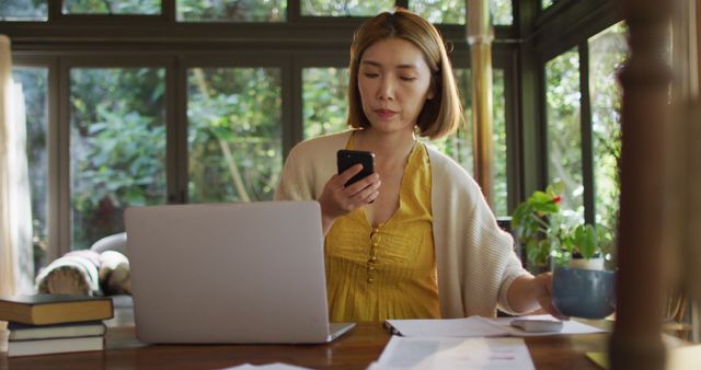 This image depicts an Asian woman working from home using a laptop and smartphone at a wooden table. Dressed in a yellow top and white cardigan, she is calmly multitasking during a workday. There is greenery visible through large windows, which bring in natural light, enhancing the serene ambiance. Ideal for articles or blogs about remote work, productivity tips, professional women's lifestyle, and balancing work and home life.