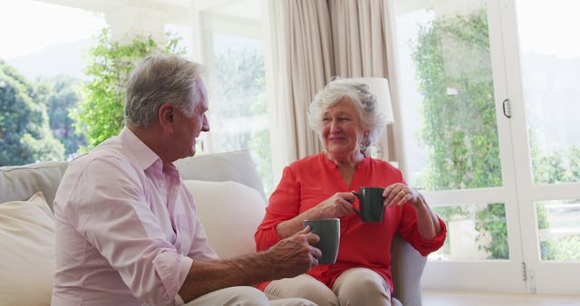 Happy caucasian senior couple sitting in sunny living room talking and drinking cups of coffee. at home in isolation during quarantine lockdown.
