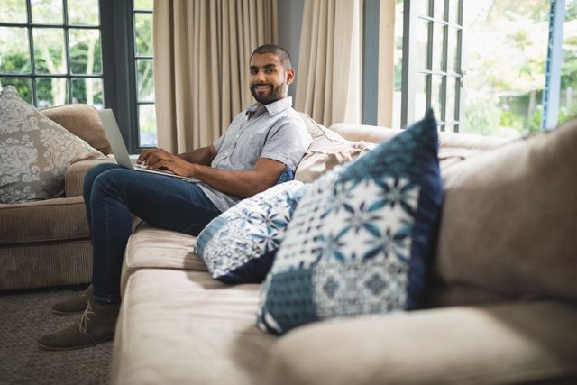 Man sitting on couch using laptop, smiling and looking relaxed. Ideal for illustrating remote work, home office setups, modern lifestyle, and technology use in everyday life.