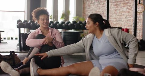 Female workout partners stretching and chatting on gym floor near dumbbells and brick wall