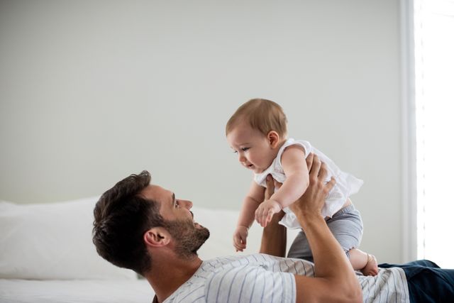 Father lying on bed holding baby up, both smiling and enjoying moment. Perfect for family-oriented advertisements, parenting blogs, and articles about fatherhood and bonding with children.