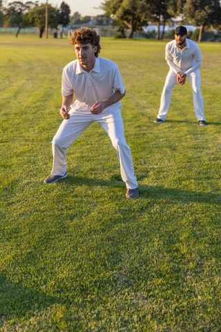 Two fielders crouching in white cricket kit on sunlit grass preparing for catch