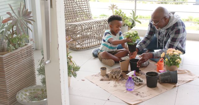 Grandfather and grandson engaging in gardening activity on porch, planting flowers in potted soil. Ideal for promoting family bonding, togetherness, and multigenerational relationships. Suitable for themes around gardening, home activities, summertime outdoors, and teaching moments.