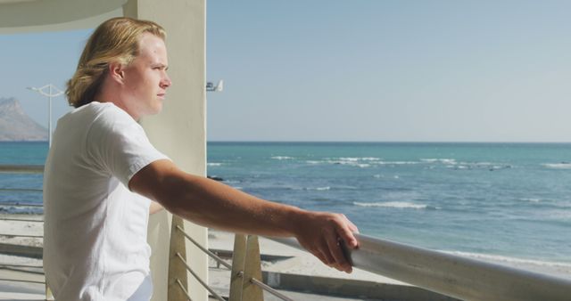 This serene image shows a young man standing on a balcony, gazing thoughtfully at the ocean on a bright sunny day. Perfect for illustrating concepts of relaxation, contemplation, leisure, mental well-being, outdoor activities, coastal living, and serene vacation scenes.
