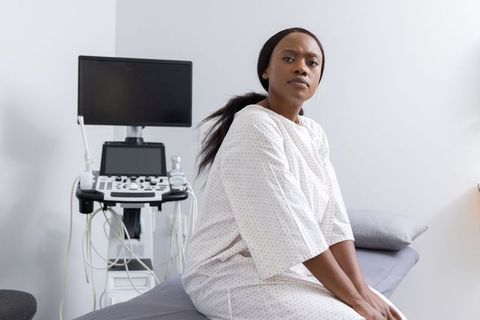African woman in medical gown sitting on exam table watching ultrasound monitor with concerned expre