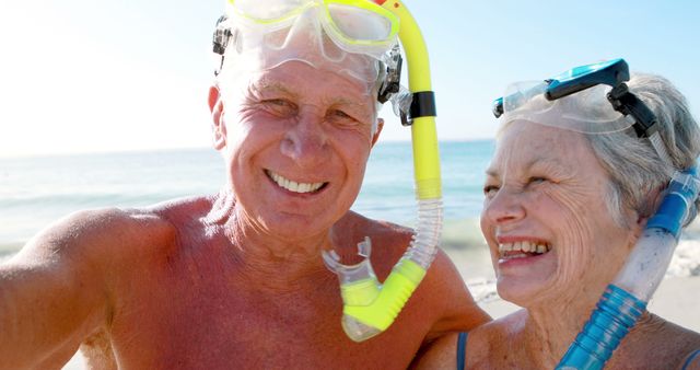 This stock photo shows an elderly couple in snorkeling gear at a beach. Both are smiling and looking happy, suggesting a joyful, adventurous lifestyle. Perfect for use in articles or advertisements about senior travel, active aging, retirement activities, or vacation destinations. It evokes themes of happiness, health, and the joys of an adventurous spirit in later years.