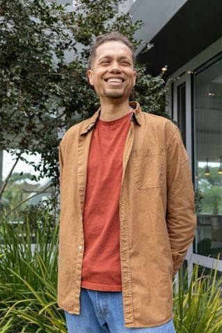 Smiling man in his 30s standing in courtyard wearing rust t-shirt and tan overshirt