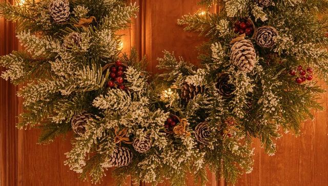 Hanging flocked evergreen wreath with pine cones red berries warm lights on wood door