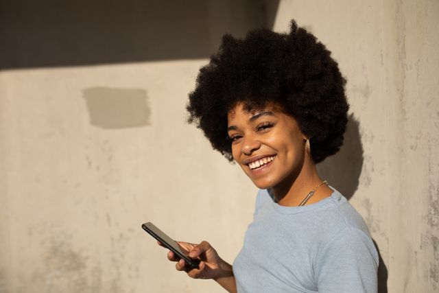 Portrait of a happy biracial woman enjoying free time in a city on a sunny day, using a smartphone, smiling leaning against a wall, wearing blue top.