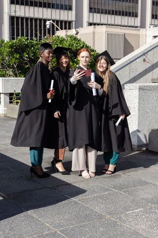 Diverse female graduates taking selfie on urban campus plaza wearing caps and gowns