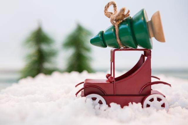 Toy car carrying a Christmas tree on snowy ground. Ideal for holiday greeting cards, festive decorations, winter-themed marketing materials, and Christmas advertisements.