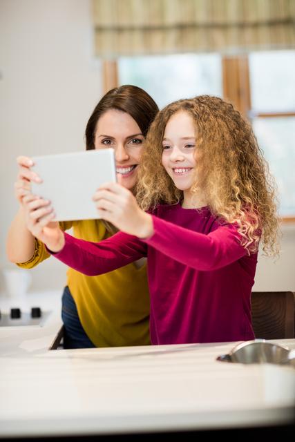 Mother and daughter smiling while taking a selfie with a digital tablet in a modern kitchen. Perfect for themes related to family bonding, technology use in everyday life, parent-child relationships, and happy home moments.