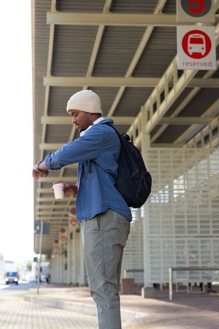 African american commuter checking watch while holding takeaway coffee at bus shelter