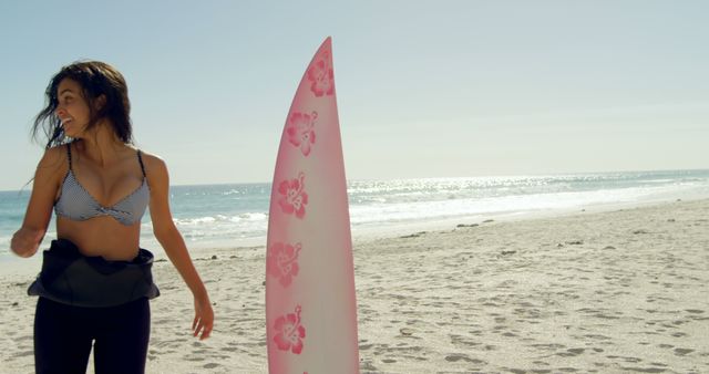 Young biracial woman stands by a surfboard on the beach, with copy space. She enjoys a sunny day at the seaside, ready for a surfing session.