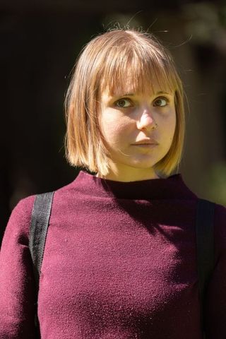 Young woman standing in park wearing maroon sweater and backpack, short bob, pensive portrait