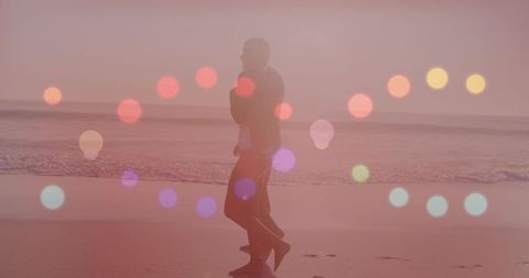 Couple walking along pastel beach at sunset with dreamy bokeh orbs and soft silhouettes