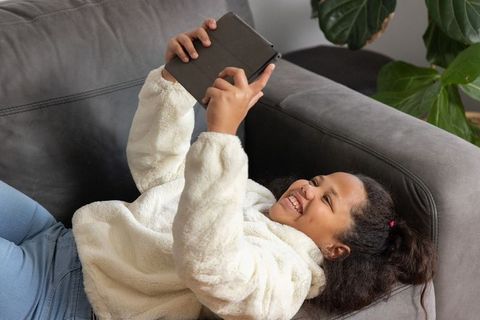 African american girl reclining on sofa using tablet, smiling in cozy white fleece jacket