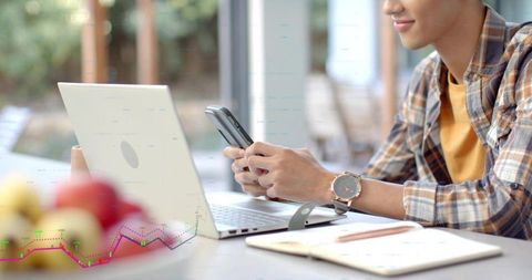 Young man checking smartphone while working remotely on laptop with notebook
