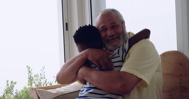 A senior man is hugging his grandson near a large window with plants visible outside. This scene of love and family bonding is perfect for use in family-focused content, advertisements celebrating family ties, or blog posts about the importance of family relationships.