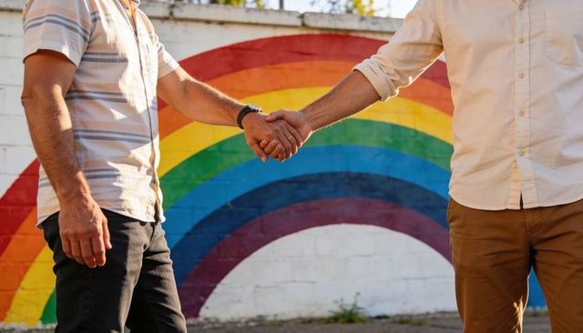 Shaking hands between two men in front of rainbow mural symbolizing unity, pride and inclusion
