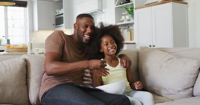 Father and daughter are sitting on a couch in a cozy living room, both smiling and enjoying a television show together. This image is perfect for depicting family bonding, parental love, and quality family time. It can be used in advertisements, articles, and blog posts related to family life, television, parenting tips, or living room decor ideas.