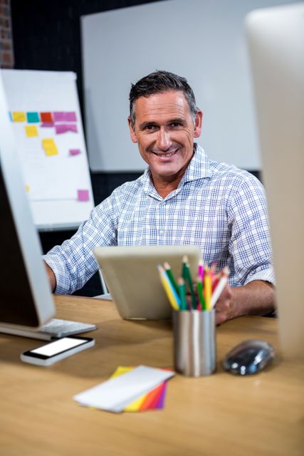 Middle-aged businessman smiling while using a digital tablet in a modern office. Ideal for themes related to business, technology, professional work environments, productivity, and modern office settings. Useful for illustrating articles, blog posts, and advertisements about workplace happiness, digital tools in business, and professional success.