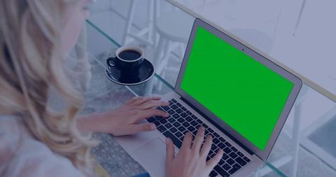 Woman typing on green-screen laptop in modern cafe workspace with coffee and glass table