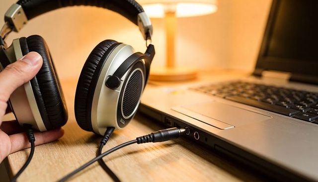 Hand holding over-ear headphones plugging 3.5mm jack into laptop on wooden desk
