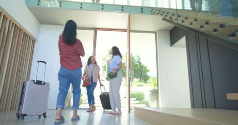Women entering sunlit modern home carrying rolling luggage and handbags while chatting