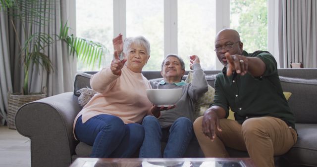 Senior friends sitting on a couch in a living room engaging in an enthusiastic TV watching session. This can be used for concepts related to socializing among elderly people, home entertainment for seniors, retirement lifestyle, and happiness in old age.