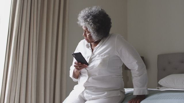 Elderly African American woman sitting on bed, using smartphone. Ideal for illustrating technology use among seniors, modern lifestyle in retirement, and elderly staying connected. Suitable for content related to senior lifestyle, tech adoption, home settings, and day-to-day activities during retirement.